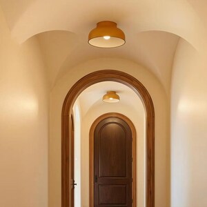 An arched hallway with a series of doorways and ceiling lights. The walls are painted a light beige, and the doorways are framed with dark wood. Two yellow ceiling lights are visible in the image.