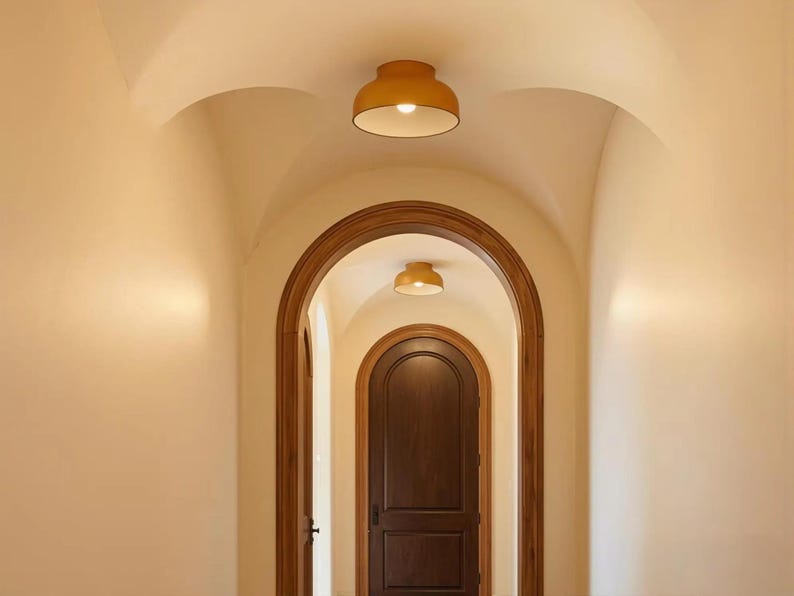 An arched hallway with a series of doorways and ceiling lights. The walls are painted a light beige, and the doorways are framed with dark wood. Two yellow ceiling lights are visible in the image.