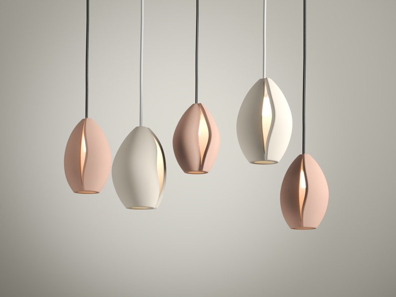 May include: Five modern pendant lights in a row, each with an oval shape and a vertical slit revealing a warm glow. The lights are in shades of pink and white, suspended by black and silver cords against a neutral background.