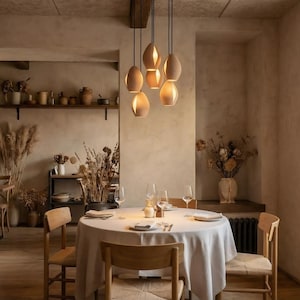 May include: A dining room with a round table set for a meal, surrounded by wooden chairs. Above the table, six pendant lights hang from the ceiling. The room has a neutral color palette with dried flowers and decorative items.