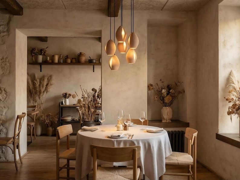 May include: A dining room with a round table set for a meal, surrounded by wooden chairs. Above the table, six pendant lights hang from the ceiling. The room has a neutral color palette with dried flowers and decorative items.