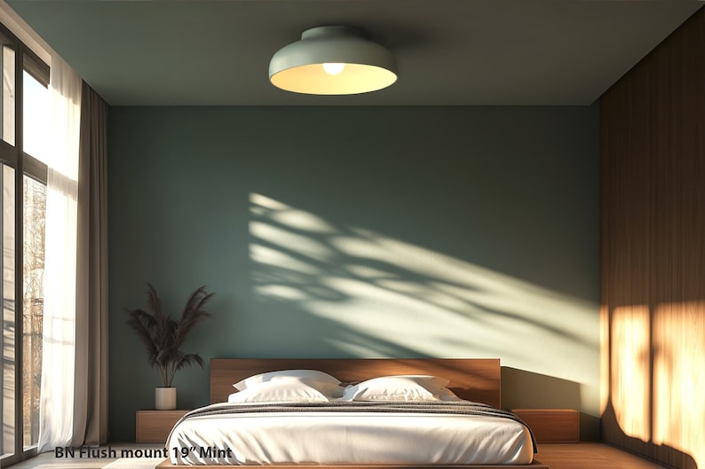 A mint-colored flush mount ceiling light illuminates a bedroom with a wooden bed frame and nightstands. The room features a green wall, a large window with sheer curtains, and a plant in a white pot. The text "BN Flush mount 19" Mint" is visible.