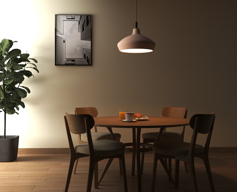 May include: A round wooden dining table with four matching chairs featuring upholstered seats. A pendant light hangs above the table, which holds a glass of orange juice and a small plate. A potted plant stands to the left, and a framed print hangs on the wall.