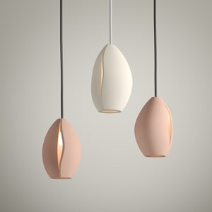 May include: Three modern pendant lights in a row, each with an elongated, organic shape. Two are a soft pink, and one is white. Each light has a vertical slit revealing a warm glow. Black cords suspend them from above.