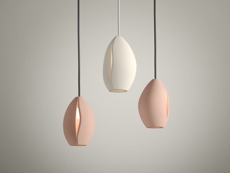 May include: Three modern pendant lights in a row, each with an elongated, organic shape. Two are a soft pink, and one is white. Each light has a vertical slit revealing a warm glow. Black cords suspend them from above.