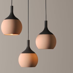 May include: Three contemporary pendant lights. Each light has a black top and a rounded, peach-coloured base. The lights are suspended from black cords against a neutral backdrop, with one light emitting a warm glow.