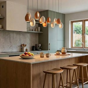 May include: A modern kitchen with a light wood island and matching stools. Above the island hang several pendant lights with brown and white teardrop-shaped shades. The kitchen features sage green cabinets and a window overlooking a backyard.