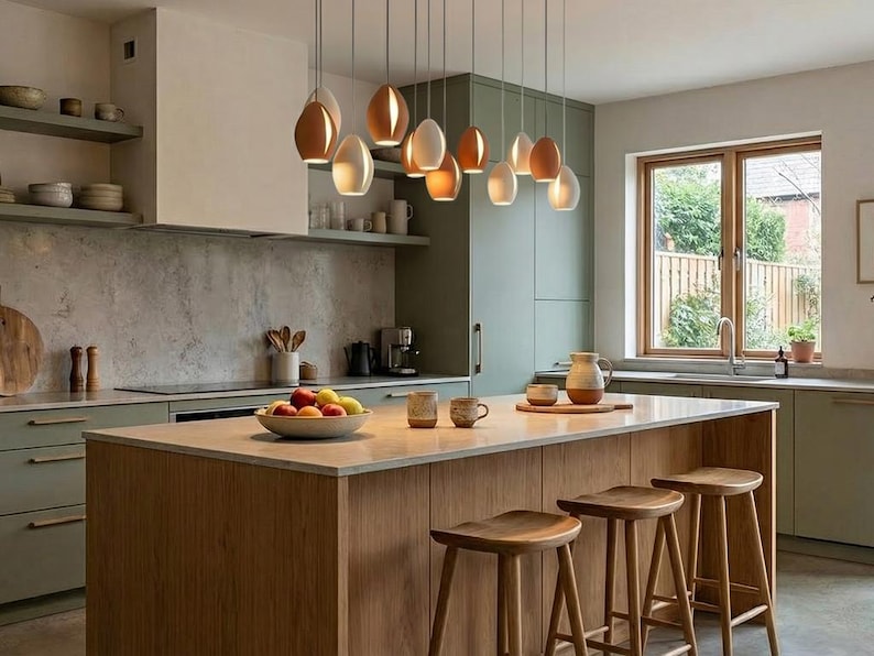 May include: A modern kitchen with a light wood island and matching stools. Above the island hang several pendant lights with brown and white teardrop-shaped shades. The kitchen features sage green cabinets and a window overlooking a backyard.
