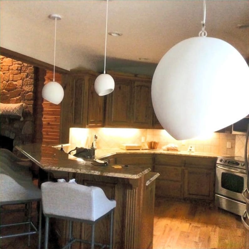 May include: A kitchen with a granite countertop and wooden cabinets. Three white spherical pendant lights hang from the ceiling. A stainless steel oven is visible on the right side of the image. The kitchen has a warm, inviting atmosphere.