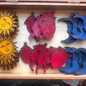 May include: A wooden box containing stacks of laser-cut wooden shapes. The collection includes yellow suns with faces, red anatomical hearts pierced by arrows, and blue crescent moons with faces. The items are arranged in rows within the box.