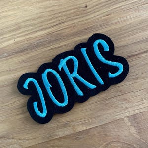 Könnte beinhalten: Ein schwarzer, gestickter Aufnäher mit dem Namen "JORIS" in hellblauer Schreibschrift. Der Aufnäher hat einen wellenförmigen Rand und liegt auf einer Holzoberfläche.