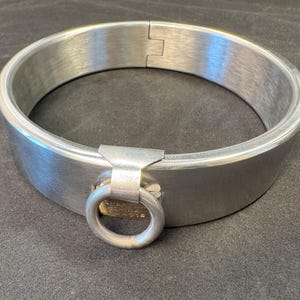 May include: A circular silver-toned metal collar featuring a brushed finish, a hinged closure mechanism, and a central metal ring attached to a small rectangular plate. The design is simple and industrial, with a smooth, polished interior surface.
