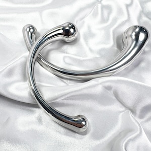 Puede incluir: Dos objetos metálicos pulidos de color plateado con un diseño curvo en forma de U. Cada pieza tiene un extremo redondeado y bulboso y una superficie lisa y reflectante. Los objetos se muestran sobre una tela blanca satinada.