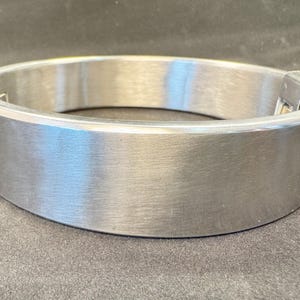 May include: A wide, circular metal collar with a brushed silver finish. The band features a smooth, continuous surface with a visible hinged locking mechanism on one side, constructed from a durable, polished metallic material with a flat profile.