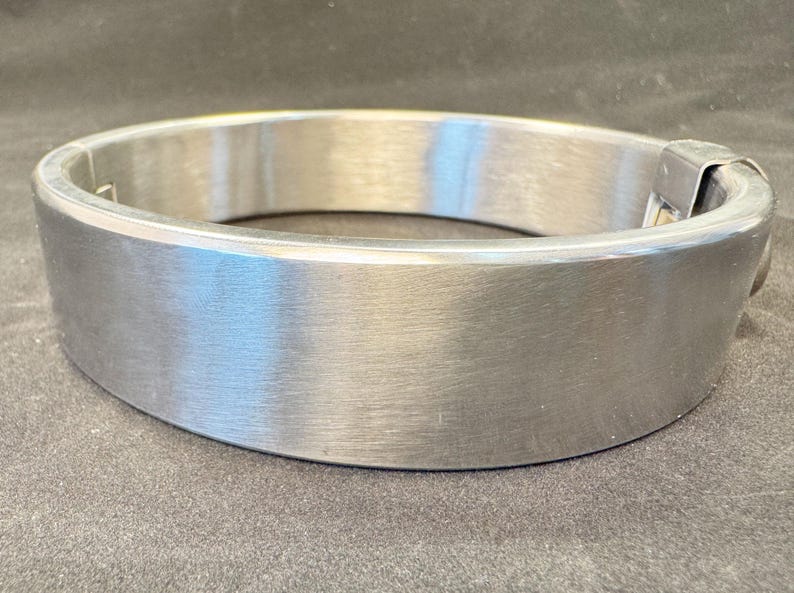 May include: A wide, circular metal collar with a brushed silver finish. The band features a smooth, continuous surface with a visible hinged locking mechanism on one side, constructed from a durable, polished metallic material with a flat profile.