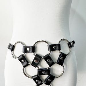May include: Black leather and silver metal body harness. The harness features a series of interconnected silver rings with black leather straps and silver rivets. The design is centered around the waist and hips.