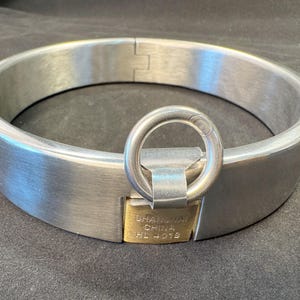 May include: A circular silver-toned metal collar featuring a brushed finish and a hinged closure. A central brass-colored padlock is integrated into the front, displaying the text SHANGHAI CHINA HL 401B, and includes a circular metal ring attachment.