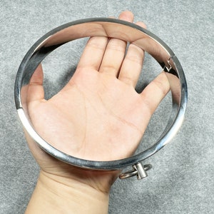 May include: A silver metal choker with a hinged clasp. The choker is shown on a hand, with the opening of the choker facing the camera.