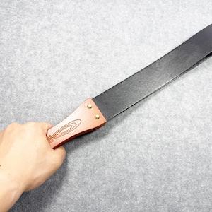 May include: A straight razor strop with a black leather surface and a wooden handle. The handle is brown with a decorative design and brass rivets. The strop is held against a gray background.