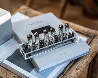 May include: A digital clock with six Nixie tubes displaying green numbers. The clock is mounted on a clear acrylic base and sits on top of two books. A concrete lamp base is visible in the background.