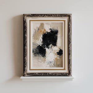 May include: Abstract art painting in a decorative silver and gold frame. The artwork features a textured composition of black, white, and tan brushstrokes. The framed art is displayed on a white shelf against a white wall.