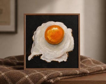 Puede incluir: Un cuadro cuadrado enmarcado que muestra un huevo frito con una yema de color naranja brillante y clara blanca sobre un fondo negro sólido. La obra de arte está encerrada en un marco de madera fino de color marrón claro.