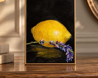 Puede incluir: Una pintura al óleo en un marco dorado fino que muestra un limón amarillo brillante sobre un fondo negro oscuro. Una rama de flores de lavanda moradas está colocada horizontalmente sobre la parte inferior del limón, creando un contraste.