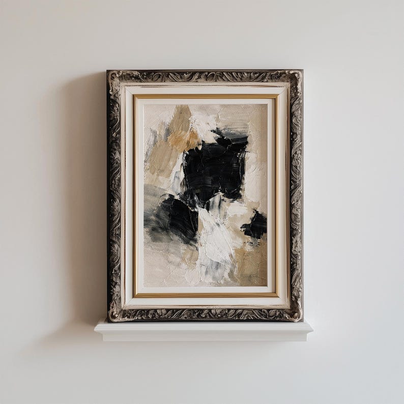May include: Abstract art painting in a decorative silver and gold frame. The artwork features a textured composition of black, white, and tan brushstrokes. The framed art is displayed on a white shelf against a white wall.