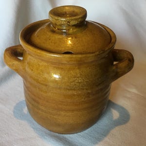 Puede incluir: Una vasija de cerámica color amarillo mostaza con tapa a juego y dos asas laterales. La vasija presenta un cuerpo cilíndrico con sutiles estrías horizontales y una pequeña muesca en el borde de la tapa, con un acabado de esmalte brillante.