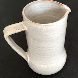 Puede incluir: Jarra de cerámica artesanal de color blanquecino, con asa curva y pico vertedor. La jarra tiene una textura moteada y una forma rústica y orgánica. El interior y el borde tienen un tono marrón rojizo. La jarra mide unos 18 cm de altura.