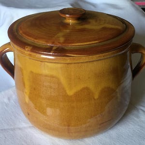 Puede incluir: Una olla de cerámica vidriada de forma redonda con dos asas laterales y una tapa a juego. El exterior presenta un color base amarillo mostaza cálido con patrones de esmalte marrón oscuro que gotean desde el borde hacia la base.
