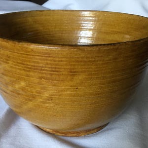 Può includere: Una ciotola in ceramica rotonda e profonda con una smaltatura lucida color giallo senape. La superficie esterna presenta sottili striature orizzontali che circondano il corpo, creando un aspetto strutturato. La ciotola ha un'apertura ampia e una base leggermente affusolata.