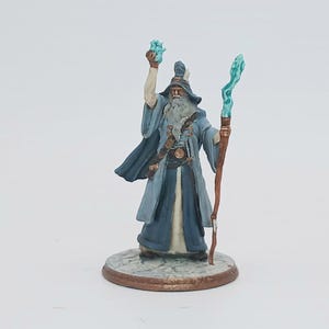 May include: A miniature figurine of a wizard in a long blue robe and hat, holding a glowing blue orb in one hand and a staff with a blue crystal at the top in the other. The wizard has a long white beard and is standing on a round base.
