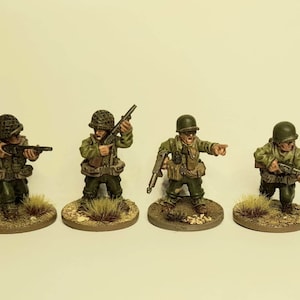 May include: Four painted miniature figures of soldiers wearing green uniforms and helmets. Each figure is holding a rifle and standing on a small brown base with green grass.