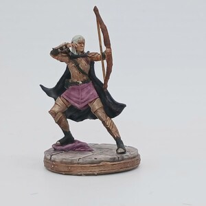 May include: A miniature figurine of an archer in a combat stance, poised to fire an arrow. The figure is painted with gold, black, and purple accents. The archer is standing on a circular, stone-like base. The bow is brown.
