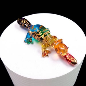 Puede incluir: Una figurita de vidrio colorida de un ornitorrinco con un esquema de color negro, azul, verde, amarillo, naranja y rojo. El ornitorrinco tiene detalles dorados y un pico largo y delgado.