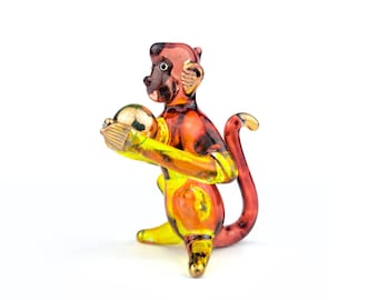 May include: A glass monkey figurine in red, orange, and yellow hues, holding a golden ball. The monkey has a curled tail. The figurine is set against a white background.