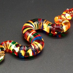 Könnte beinhalten: Eine farbige Glasschlangenfigur mit roten, gelben, grünen und blauen Streifen. Die Schlange ist S-förmig gekrümmt und hat einen goldfarbenen Schwanz.