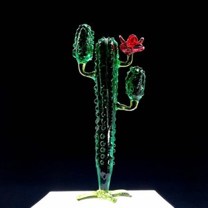 May include: A decorative glass cactus sculpture. The cactus is a vibrant green with a textured surface and a red flower. The sculpture has a detailed design and is set against a dark background.
