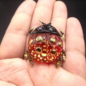 Puede incluir: Una figura de mariquita roja y dorada con cabeza negra y detalles dorados. La mariquita tiene un cuerpo rojo transparente con lunares dorados y patas doradas. La figura se sostiene en una mano.