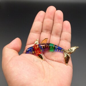 May include: A colourful glass dolphin figurine with gold accents. The dolphin is swimming with its tail up and its head tilted down. The dolphin is decorated with red, blue, green, and gold stripes.