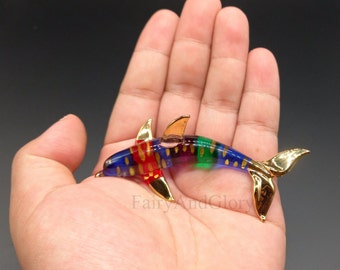 Puede incluir: Una figurita de delfín de vidrio colorido con detalles dorados. El delfín está nadando con la cola hacia arriba y la cabeza inclinada hacia abajo. El delfín está decorado con rayas rojas, azules, verdes y doradas.