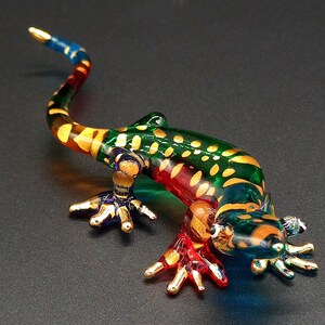 May include: A colorful glass lizard figurine. The lizard has a green body with yellow spots, a red belly, and a blue and gold tail. The feet are gold. The figurine is on a dark surface.
