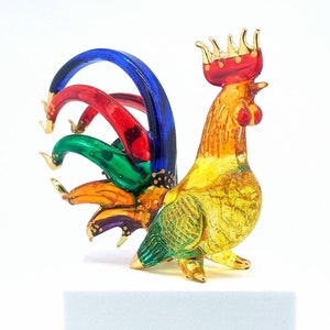 May include: A colorful glass rooster figurine with a multi-colored tail and a gold crown. The rooster is yellow, orange, green, red, and blue.