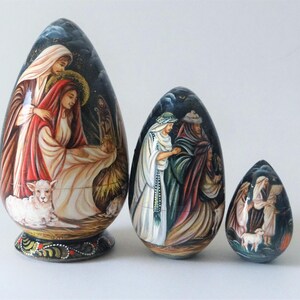 May include: Three painted wooden egg ornaments depicting religious scenes. The largest egg features figures in robes, a lamb, and a starry background. The other two eggs show similar scenes with different figures and settings. The eggs are painted in various colors.