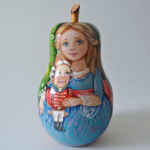 May include: A hand-painted wooden nesting doll, or matryoshka, depicting a girl in a blue dress holding a toy soldier. The doll is decorated with green, blue, red, and white paint.
