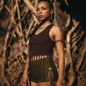 May include: A woman wearing a brown tank top with fringe detailing and olive green shorts with cut-out accents. The background features a complex network of tree roots, creating a natural, earthy aesthetic. The outfit is accessorized with silver jewelry.