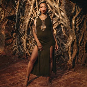 May include: An olive green maxi dress with a cut-out design on the bodice and high slits on both sides. The dress is worn in front of a backdrop of tree roots. The model is wearing silver jewelry.