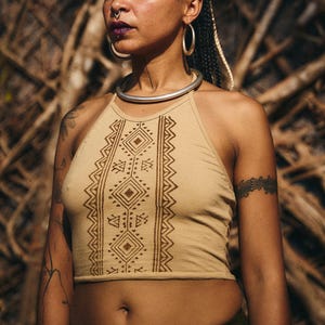 Peut inclure: Haut de bikini beige avec un motif tribal géométrique marron. Le haut présente une encolure haute et une silhouette ajustée. La personne porte un ras du cou argenté et de grandes créoles. L'arrière-plan est une vue floue de branches d'arbres.