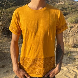 Op de afbeelding: Een mosterdgeel T-shirt met korte mouwen en een donkerbruin geometrisch patroon langs de onderzoom en op de randen van beide mouwen. Het shirt heeft een ronde halslijn en een rechte zoom, gemaakt van een effen gekleurde stof.